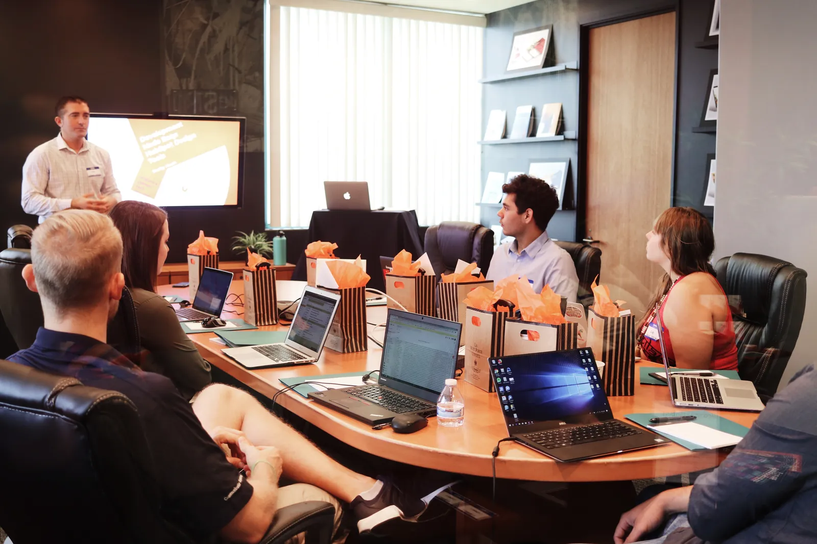 A presenter leading a meeting with a team working on laptops around a long table